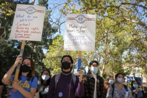 UCLA students carry pickets reading "Make Your Own Sign. Support UC Lecturers" and "What do we Need? Contract, Rehiring Rights, Living Wage, Health Care" at a UC-AFT rally.