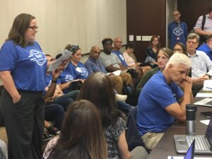 Members of the Unit 18 bargaining team are seated at a long desk with laptops open. Behind them, members in blue union t-shirts, some sitting, some standing, observe. On the left, one member, standing, addresses the university's negotiators (not pictured).