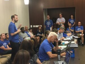 Members of the Unit 18 bargaining team are seated at a long desk with laptops open. Behind them, members in blue union t-shirts, some sitting, some standing, observe. On the left, one member, standing, addresses the university's negotiators (not pictured).