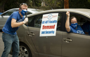 Members participate in a car caravan action at UCLA in 2020. On the left, a members of UC-AFT, in a union t-shirt and CFT mask, speaks on a microphone and on the right, a member in a union t-shirt and CFT mask raises her fist through the window of her car. The sign on the car's exterior reads "UC-AFT Faculty Demand Job Security"