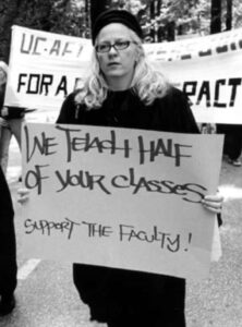 Black and white image shows a march of faculty, staff, and students at UCSC during UC-AFT's strike in 2003. Women in center of the frame is wearing black and glasses and holding a handmade sign that reads "We Teach Hard of Your Classes - Support the Faculty!". She's walking in front of a banner (not legible).