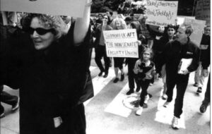 Black and white image shows a march of faculty, staff, and students at UCSC during UC-AFT's strike in 2003. On the left side of the frame is a woman carrying a sign (not pictured), in the center, a woman holds a sign reading "Support UC-AFT The Non Senate Faculty Union" - to her right, a student holds a sign reading "Real Classes- Real Faculty! Support Faculty"