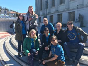 A group of UC Berkeley librarians sitting on the steps of Bancroft Library in 2018. They are wearing blue t-shirts that read "Support Librarians - University of California" with the silhouette of the state of California. Most are wearing sunglasses, one is carrying a baby.