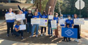 Demonstration teachers at the UCLA Lab School, many wearing blue UC_AFT t-shirts, post for a picture during their strike in 2023. Most are holding home made signs, which read "Teachers support students - please support us!", "Together we can make the UCLA Lab School better", "Teaching conditions are learning conditions", Teachers love the Lab School!", and "Teachers advocating for excellent teaching and learning conditions". One student (approximately 7 years old) holds a blue UC-AFT sign.