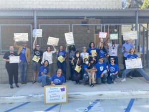 Demonstration teachers at the UCLA Lab School, many wearing blue UC_AFT t-shirts, post for a picture during their strike in 2023. Most are holding home made signs, which read "Teachers support students - please support us!", "Together we can make the UCLA Lab School better", "Teaching conditions are learning conditions", Teachers love the Lab School!", and "Teachers advocating for excellent teaching and learning conditions". They are joined by parents and supporters raising their fist and cheering.