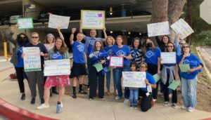 Demonstration teachers at the UCLA Lab School, many wearing blue UC_AFT t-shirts, post for a picture during their strike in 2023. Most are holding home made signs, which read "Teachers support students - please support us!", "Together we can make the UCLA Lab School better", "Teaching conditions are learning conditions", Teachers love the Lab School!", and "Teachers advocating for excellent teaching and learning conditions". They are joined by parents and supporters raising their fist and cheering.