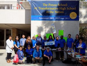 A group of teachers outside the Preuss School, most wearing blue t-shirts. Some are holding signs that read "Contract Now", "Out of Contract Rally!", and "Excellent Teachers Deserve an Excellent Contract".