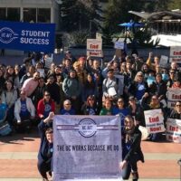 A crowd of about 100 UC Berkeley Lecturers, students, and supporters gather in Sproul plaza during the 2019-2021 contract campaign. In the foreground, two students hold a banner reading "The UC Works Because We do" with signatures of UC-AFT members. In the back, a large blue banner reads "Faculty Equity - Student Success". One student holds handmade sign reading "Students in Solidarity with teachers". Others in the crowd hold pickets reading "End Unpaid Work Now!"