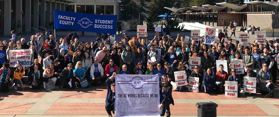 A crowd of about 100 UC Berkeley Lecturers, students, and supporters gather in Sproul plaza during the 2019-2021 contract campaign. In the foreground, two students hold a banner reading "The UC Works Because We do" with signatures of UC-AFT members. In the back, a large blue banner reads "Faculty Equity - Student Success". One student holds handmade sign reading "Students in Solidarity with teachers". Others in the crowd hold pickets reading "End Unpaid Work Now!"