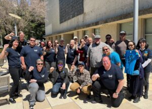 Members of the Unit 17 bargaining team and their supporters gather outside a bargaining session in April, 2024. Most are wearing dark blue t-shirts with the UC-AFT logo and raise their fists.