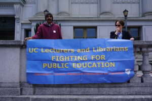 Two people hold a blue banner handing over a stone wall that reads "UC Lecturers and Librarians Fighting for Public Education - University Sundial, American Federation of Teachers"