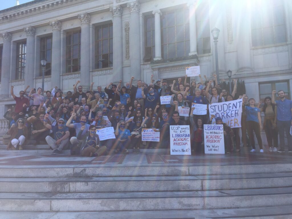 Librarians and their allies rally on the steps of the Bancroft Library at UC Berkeley in 2018. Many are wearing blue union t-shirts and holding signs, including "Student Worker Power" and "UCOP wants to end Librarian Academic Freedom - Who's Next". A ray of sunlight splashes through the top right corner.