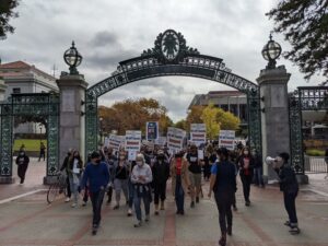 A crowd of UC Berkeley lecturers and their supporters march through the iconic Sather gate to campus in 2021. Many are wearing masks and holding picket signs reading "UC-AFT Faculty Demand Job Security".