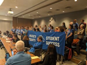 The UC-AFT bargaining team sits at the table with their laptops open during a bargaining session at the UC Office of the President in Oakland in 2019. Behind them stand members of UC-AFT, in their blue union t-shirts, holding a large blue banner that reads "Faculty Equity - Student Success". One member is reading testimony to the UC's bargaining team.