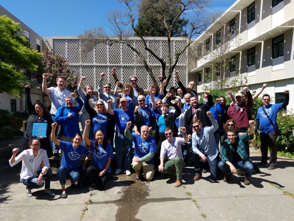 Members of the UC-AFT bargaining team and supporters raising their fists and wearing blue t-shirts outside of a bargaining session in April 2019 at UC Davis.