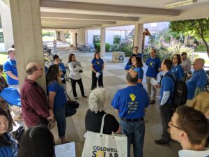 UC-AFT members stand in a circle listening to chapter chair Katie Rodger at UC Davis in 2018. Their shirts are blue, the back reads "Organize. Educate. Agitate."