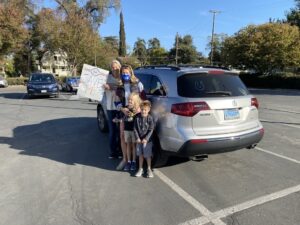 A family - two women and their two small children- participating in a car caravan action at UC Davis in 2020. One wears a blue CFT mask, the other carries a sign reading "SAVE PE".