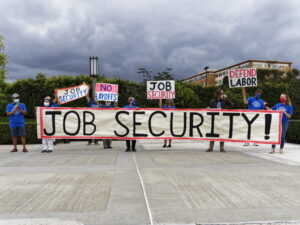 UC-AFT members in blue t-shirts standing at a safe distance during a job security action at UCI in 2020. Their banner reads "Job Security!". Some hold signs reading "Job Security" "No Layoffs" and "Defend Labor".