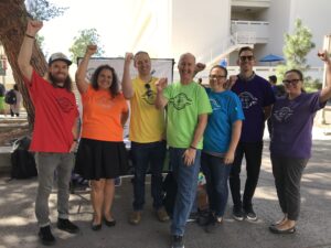 A group of lecturers in union t-shirts raising their fists in 2019. Each wears a different color t-shirt creating a rainbow (red, orange, yellow, green, blue, dark blue, and purple).