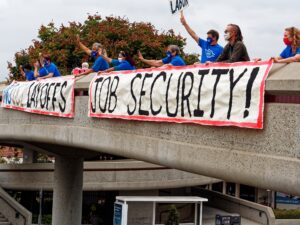 Members of UC-AFT, wearing masks and standing at a safe distance, holding a banner hanging over a pedestrian overpass at UC Irvine in 2020. Their banners read "No UCI Layoffs" and "Job Security!"