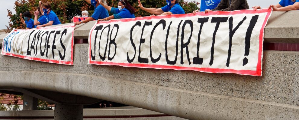 Members of UC-AFT, wearing masks and standing at a safe distance, holding a banner hanging over a pedestrian overpass at UC Irvine in 2020. Their banners read "No UCI Layoffs" and "Job Security!"