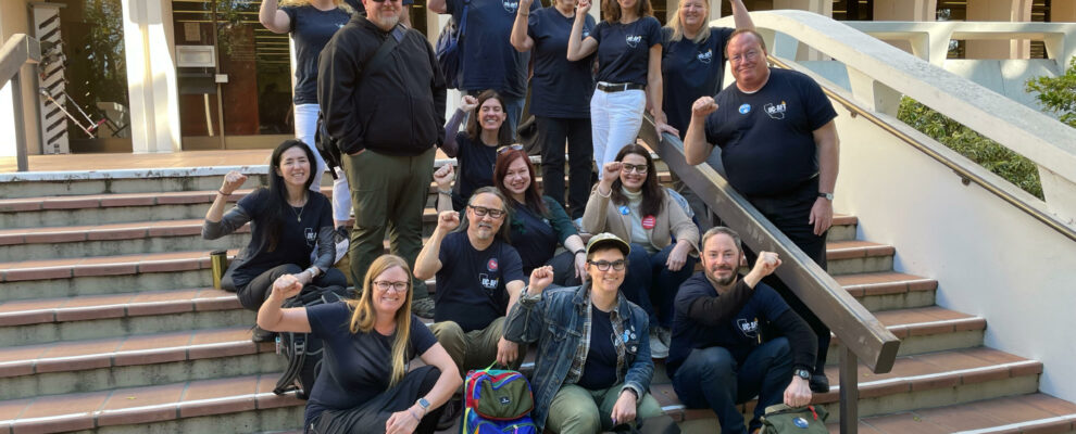 Members of the Unit 17 bargaining team and their supporters gather outside a bargaining session at UC Irvine in March, 2024. Most are wearing dark blue t-shirts with the UC-AFT logo and raise their fists.