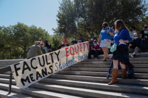 Two women, both wearing masks and blue shirts, walking down a flight of steps past a banner (fixed to the handrail) that reads "Faculty Equity Means Student Success". A crowd, most of them masked, gathers in the shade at the top of the steps.