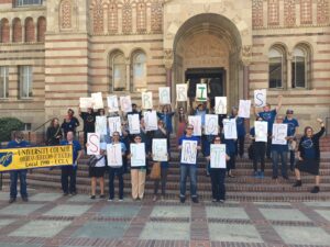 A group of librarians and supporters standing on the steps of Powell Library at UCLA in 2018. Each holds a sign with a single letter, spelling out "Librarians Will Not Be Silent." On the left, two members carry a yellow and blue banner that reads "University Council - American Federation of Teachers, local 1990 UCLA".