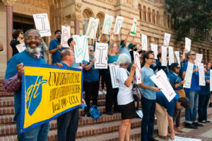 A group of librarians and supporters standing on the steps of Powell Library at UCLA in 2018. Each holds a sign with a single letter, spelling out "Librarians Will Not Be Silent." On the left, two members carry a yellow and blue banner that reads "University Council - American Federation of Teachers, local 1990 UCLA".