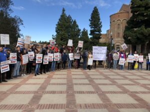 Lecturers and their students and supporters gather at the top of Janss Steps at UCLA in 2021. Many hold signs reading "End Unpaid Work Now!". Others carry a large banner reading "The UC Works Because We Do" with hundreds of members signatures.