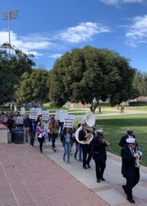 A marching band, wearing uniforms and white hats and carrying trumpets and a large tuba, lead a march of UCLA members and students at UCLA in 2021. Some carry signs reading "UC-AFT Faculty Demand Job Security"