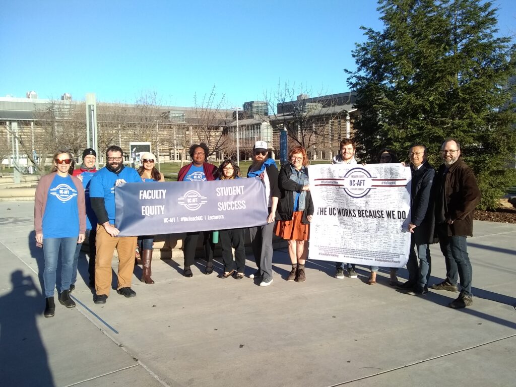 Lecturers carrying banners, many wearing blue UC-AFT t-shirts, at UC Merced in 2019. The banner on the right reads "The UC Works Because We do" with hundreds of members' signatures. The banner on the left reads "Faculty Equity - Student Success."