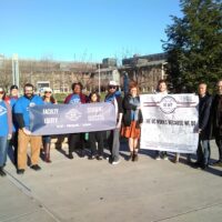 Lecturers carrying banners, many wearing blue UC-AFT t-shirts, at UC Merced in 2019. The banner on the right reads "The UC Works Because We do" with hundreds of members' signatures. The banner on the left reads "Faculty Equity - Student Success."