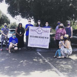 Masked members of the UC Riverside chapter photographed during a car caravan action in 2020. They are holding a banner that reads "The UC Works Because We Do" with hundreds of member signatures.