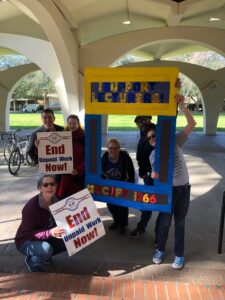 A group of lecturers from UC Riverside pose during an action in 2019. Their signs read "End Unpaid Work Now!". Others hold a handmade photo frame that reads "Support Lecturers - UC-AFT 1966"