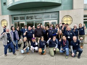 Members of the Unit 17 bargaining team and their supporters gather outside a bargaining session at UC Riverside in 2024. Most are wearing dark blue t-shirts with the UC-AFT logo and raise their fists.