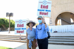 A young couple holding picket signs reading "End Unpaid Work Now!" at UCSB in 2020. The woman has a sleeping baby nested on her chest in a wrap.