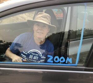 A member of UC-AFT in her blue union t-shirt participates in a car caravan action in 2020. She is sitting in the passenger seat of her car, which she has decorated with blue paint to mimic the square effect on zoom (thin outline with zoom written in the corner).