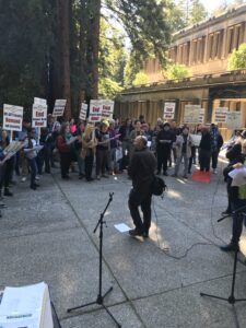 UCAFT member Josh Brahinnsky addresses a crowd carrying picket signs in the center of a redwood grove at UC Santa Cruz in 2021. The picket signs read "End Unpaid Work now!" and "UC-AFT Faculty Demand Job Security"