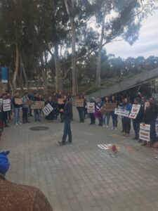 A male member of UC-AFT addresses a crowd of students and supporters at a rally at UC San Diego in 2021. The rally is near a grove of eucalyptus trees. Those listening hold printed signs reading "End Unpaid Work Now!" and "UC-AFT Faculty Demand Job Security." Others hold handmade signs, including "UC for the Many" and "We stand with our teachers!"