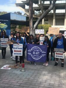 Members of UC-AFT, some in blue union t-shirts, stand together at a rally in 2021. They carry a UC-AFT banner that reads "Faculty Equity-Student Success," printed signs reading "UC-AFT Faculty Demand Job Security," and handmade signs reading "Reasonable Workload," "We Stand for Teaching" and "EVC Simmons - Practice What you Preach"