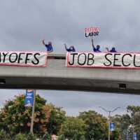 Members of UC-AFT, wearing masks and standing at a safe distance, holding a banner hanging over a pedestrian overpass at UC Irvine in 2020. Their banners read "No UCI Layoffs" and "Job Security!" One member holds a picket sign that reads "Defend Labor"