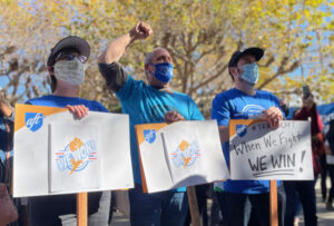 Three masked UC-AFT members holding signs while listening to a speaker at the UC-AFT victory rally in 2021. Their signs read "We Won!" and "When we fight we win!". One is raising his fist.