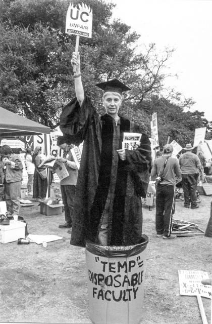 In this black and white image, a lecturer, wearing graduation regalia, stands in a trash can, holding a book and raising a small sign in the style of the statue of liberty. The trash can is labelled "temp disposable faculty" - the book reads "Respect" and her sign reads "UC Unfair."