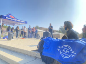 On the right, a pile of UC-AFT blue t-shirts in a box, which a student seated behind it. On the left, a blue tent labelled "Central Coast Labor Council" with a few people standing under it. In the center, a speaker on a microphone addresses the crowd. Sun splash makes the image slightly blurry/out of focus.