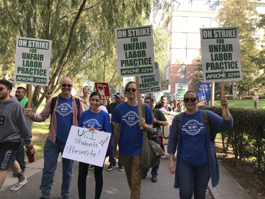 Lecturers in blue union t-shirts carrying green and white picket signs reading "On strike for Unfair Labor practice - AFSCME 3299"