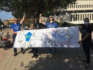 Four people, some in medical masks, some raising their fists, hold a homemade banner with a cartoon anteater wearing a UCI sweater. Out of the anteater's nose shoots an explosion-type text bubble that reads "Victory"