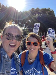 Two women in sunglasses take a selfie. Behind them, people are walking carrying picket signs reading "UAW ON STRIKE - UNFAIR LABOR PRACTICE"