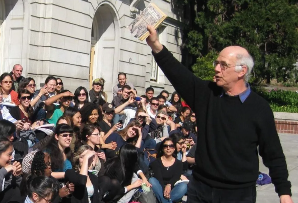 In the foreground, an older man in a black sweater stands, his hand raised in the air holding a book. He is addressing a crowd sitting on the steps behind him.