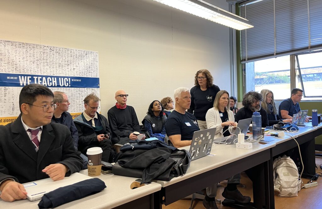 a group of lecturers sit at a long white table with a row of colleagues seated behind them. Some are wearing blue union t-shirts and have laptops open in front of them